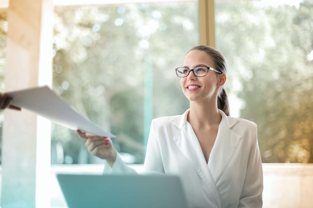 a woman in a white suit and glasses is holding a piece of paper while looking at a laptop