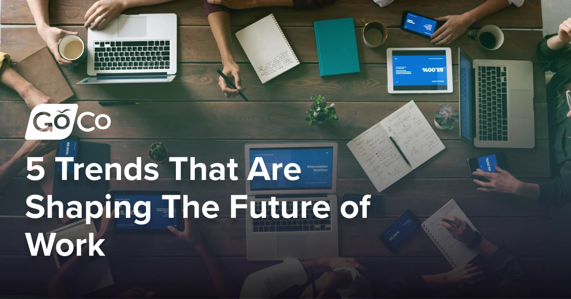 A top-down view of a table with eight people sitting at it, using laptops, tablets, phones, and notebooks.