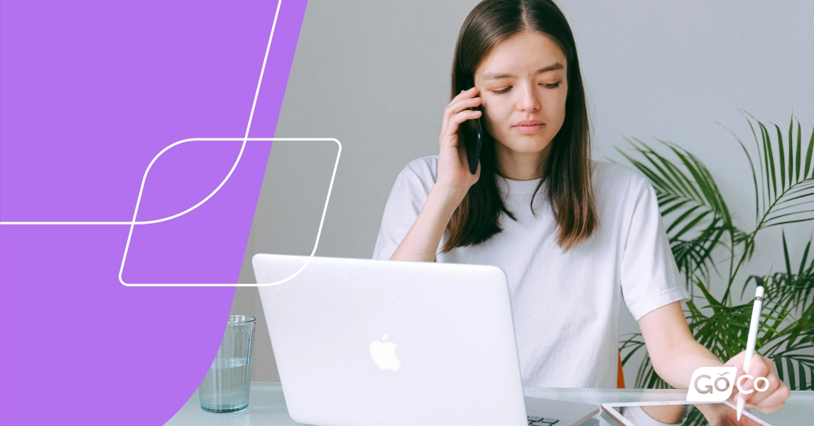 A woman sitting at a desk in front of a laptop, talking on a cell phone, and taking notes on a tablet.