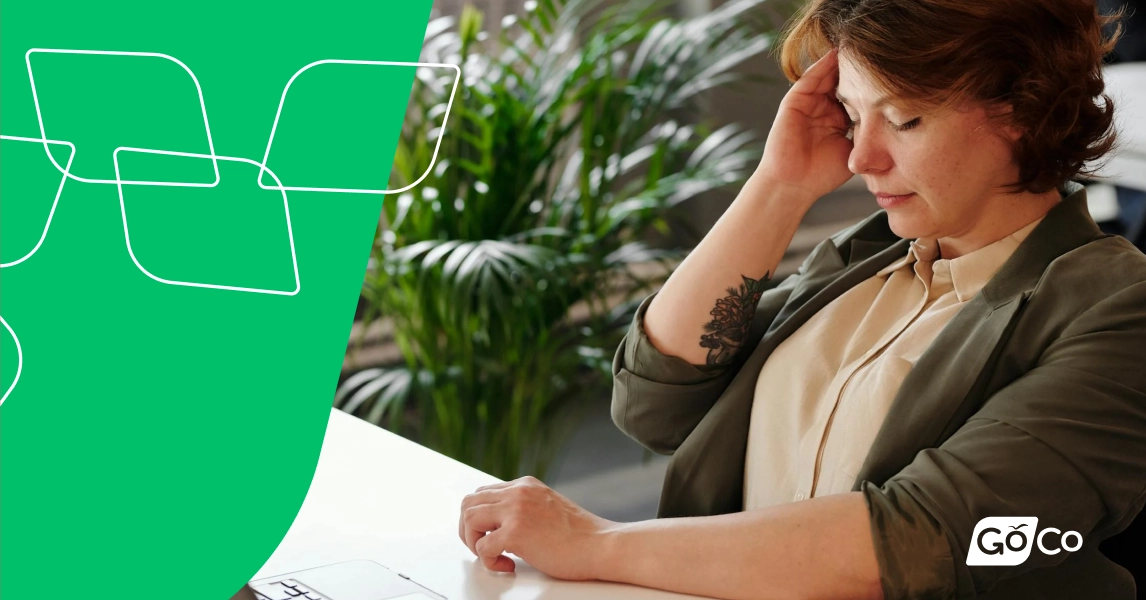 A woman sitting at a desk with her eyes closed, rubbing her temple with her right hand.