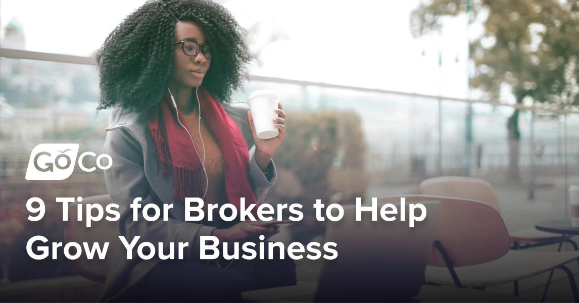 A woman sitting at an outdoor table looking at a laptop, holding a coffee in her left hand and a smartphone in her right hand.